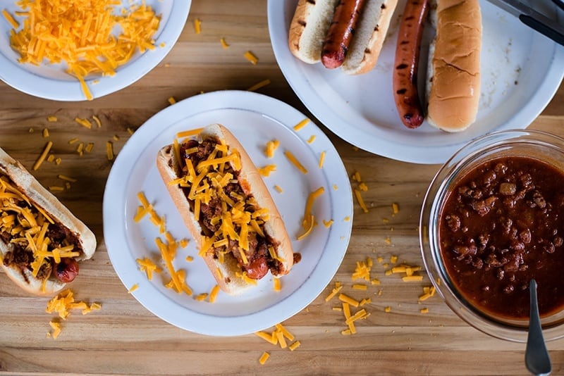 Overhead view of Chili Cheese Dogs on white plates, with a bowl of chili off to the side.