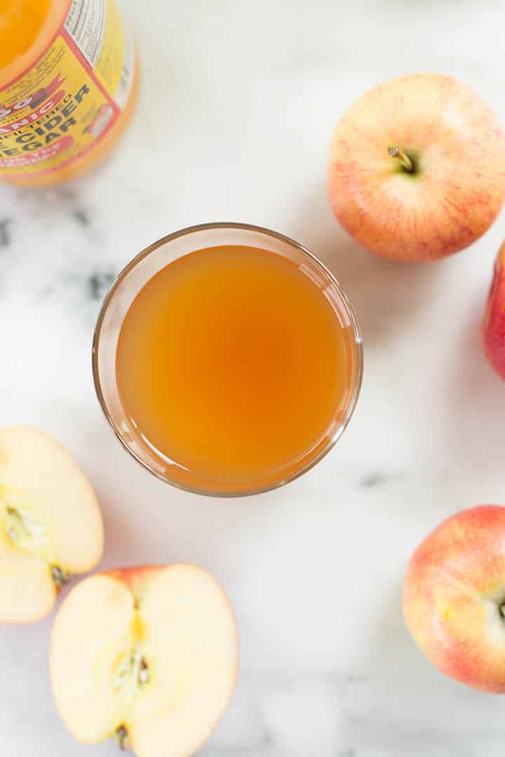 Top view of a glass of apple cider vinegar surrounded by whole apples and an apple cut in 2. 