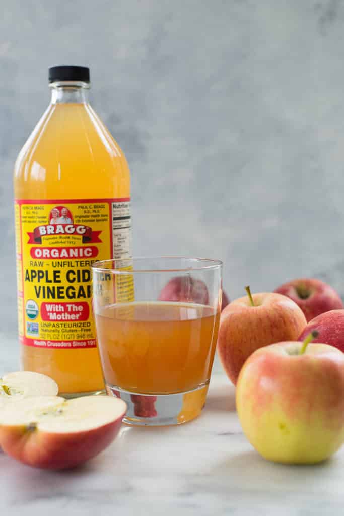 Close up image of a bottle of apple cider vinegar, a glass of water and apple cider vinegar mixture, and whole and sliced apples around the bottle and glass.