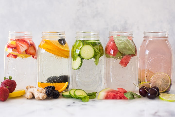 5 flavors of infused water in mason jars lined up in a row, including strawberry banana and cucumber.
