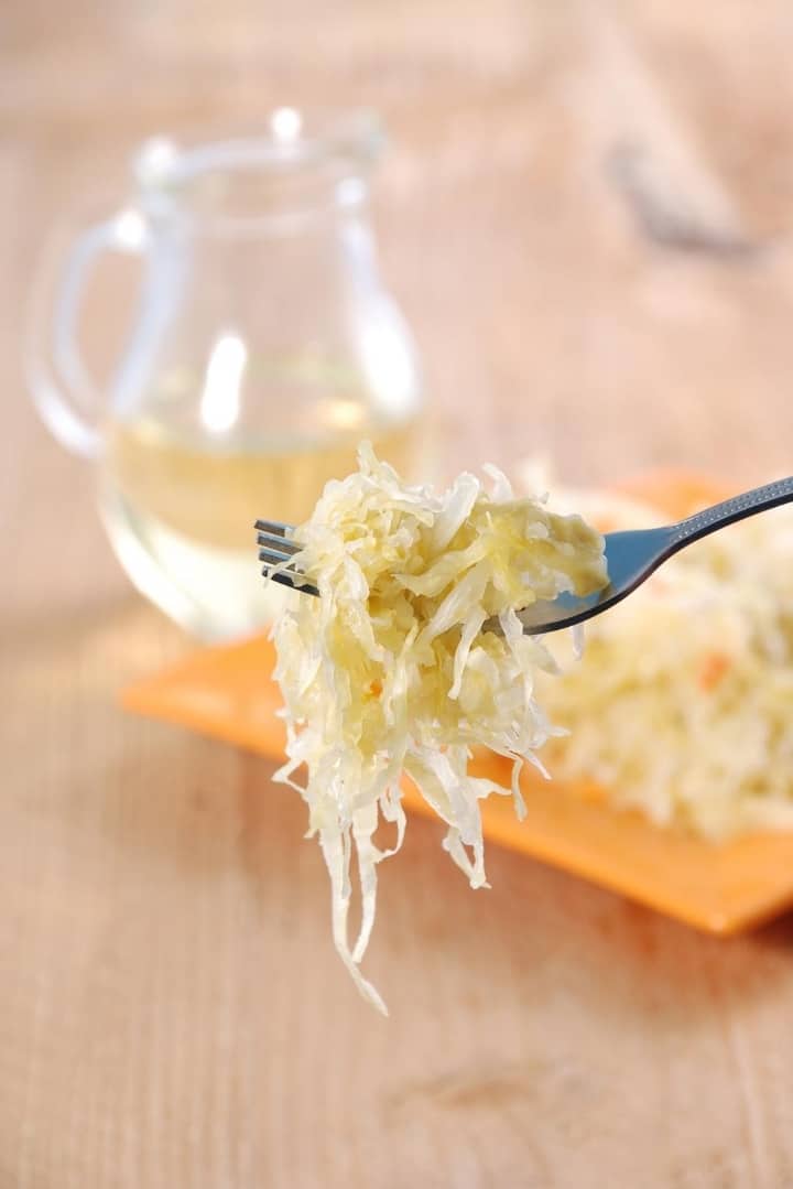Close up side view of a fork holding a portion of ready to eat sauerkraut, with a plate of sauerkraut in the background.